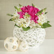 In this refined wedding arrangement, a rounded white wicker basket becomes a stage for an intimate play of colour and texture, reminiscent of the soft, polished interiors around Gerrards Cross and nearby Chalfont St Peter. At the centre, exuberant fuchsia blooms - full-headed and velvety, like peonies at their peak - gather tightly together, forming a vivid, romantic core. They are cosied in by a collar of creamy white alstroemeria, each slender petal feathered with delicate magenta and green markings that invite the eye to linger. Slim green stems and unopened buds peek here and there through the flowers, adding movement and that sense of fresh beginnings couples might feel walking through West Common on a bright spring morning. The basket itself is crafted from smooth, white-painted wicker, woven into an intricate open pattern that allows light to slip through, casting subtle shadows and making the design feel both modern and timeless. Two cream-coloured rattan spheres rest nearby, mirroring the textures of the basket and providing a serene, sculptural balance. The entire piece is bathed in soft, diffused light, which gently illuminates the blooms and highlights the crisp contrasts between fuchsia petals, ivory flowers, and clean white wicker. The mood is airy and hopeful, ideal as a refined centrepiece for a small wedding breakfast at a local Gerrards Cross venue or as a thoughtful gift delivered to a bride-to-be. Every detail, from the tight clustering of petals to the interplay of light and shadow, conveys careful craftsmanship and a love of floral design.