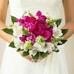 This image shows a romantic hand-tied bouquet held close, in the kind of soft daylight you might find drifting through a window near East Common in Gerrards Cross. At the centre, several large magenta peonies bloom with confident abundance, their ruffled petals folding over one another in generous curves, creating a rich cushion of colour that immediately draws attention. Framing these peonies are clusters of white alstroemeria, each bloom lightly feathered with fine burgundy or soft brown lines, lending an air of refinement and giving the arrangement a gentle rhythm. Between the open flowers, slender spikes of pale green buds and new growth reach upwards, bringing a feeling of vitality and promise, like fresh leaves appearing in spring gardens around West Common. Lush green foliage wraps around the base, grounding the composition and offering both visual balance and a subtle, leafy scent that tempers the floral sweetness. The bouquet catches natural light from one side, which softly illuminates the magenta tones of the peonies and casts subtle shadows into the folds of the petals, adding a sense of depth and quiet drama. The overall mood is one of modern elegance blended with classic romance, ideal for a bride stepping into a ceremony in Gerrards Cross or for marking an important life moment at home. Crafted with care by a local florist who understands how these tones photograph and how they feel in the hand, the bouquet speaks of celebration, tenderness, and the hopeful beginning of a new chapter.