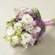 A lush hand-tied bridal bouquet, meticulously crafted by the experienced florists of Gerrards Cross, is presented atop a soft, light beige surface. Here, velvety white lisianthus blossoms with gently ruffled petals gather around creamy yellow centers, hinting at orchard blooms glimpsed along local avenues. Light pink and lavender-hued stocks are woven throughout, offering airy swirls of texture and a subtle, sweet fragrance evocative of garden evenings by the Misbourne stream. Touches of deep purple Allium 'Star of Persia' break through with their spiky, symmetrical star-formed globes, lending both contrast and European flair. Cascading foliage of rich, fresh greens and unopened lisianthus buds provides natural movement, layering the bouquet with shades seen in Gerrards Cross' lush parks. The hand-tied stems are secured by a bright fuchsia satin ribbon, finished with elegant white pearls-a signature of bridal sophistication in the area. Soft daylight enhances the bouquet's palette and detail, from the smooth lisianthus petals to the delicate, cloud-like stock. This inspiring arrangement, perfect for brides, event décor, or romantic gestures, encapsulates the refined artistry and heartfelt traditions cherished across Gerrards Cross.