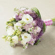 A refined, elongated bouquet lies on a pale beige, slightly textured cloth, the neutral backdrop allowing each blossom to stand out clearly, as if ready for a bridal portrait in a quiet Gerrards Cross townhouse. Dominating the arrangement are large white lisianthus, petals layered like fine silk, some fully open to reveal gentle yellow-green centres, while others remain in elegant, tapered buds that hint at blossoms still to come. Between them nestle clusters of soft pink to lilac stock flowers, each stem covered in numerous dainty, ruffled florets that build a sense of airy volume and nostalgic romance. Punctuating these pastel tones are vivid tufts of spiky purple filler blooms, similar to ornamental allium, their star-like flowers giving the bouquet lightness, movement, and a faintly whimsical character. Long, slender blades of deep green foliage weave in and out of the composition, particularly noticeable as they sweep in arcs along the right-hand side, shaping the bouquet and creating a graceful, flowing rhythm. The stems are firmly gathered and wrapped in a bright fuchsia satin ribbon, perfectly smooth and slightly lustrous, which is then refined further by a delicate line of tiny white pearls, echoing a bride's jewellery or the buttons on a wedding gown. The overall mood is romantic but not overly formal, evoking soft afternoon light filtering through homes near West Common, or a couple exchanging vows in a local Gerrards Cross venue. This is the sort of bouquet a professional florist at Flower Delivery Gerrards Cross might craft for a bride who loves gentle colour, texture, and a mix of classic and contemporary style.