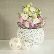 A scene of serene beauty unfolds in this handcrafted floral composition, reflecting the warmth and artistry of Gerrards Cross. Resting at the center is a round, white decorative vase, meticulously woven from painted rattan-the intricate open-work resembling artisanal baskets found at local craft fairs. Overflowing delicately from the vessel is a lush medley of soft lavender and creamy eustoma, the ruffled petals unfurling in layers suggestive of gentle spring breezes over East Common. Wisps of pale green foliage peek through, weaving freshness throughout the display. The floral abundance is grounded by vivid purple miniature blooms tucked at the base, their rich colour echoing English wildflowers and adding depth. Flanking the white vase to the left, two decorative rattan spheres-one slightly larger, their open, basket-weave construction catching daylight-accentuate the natural, tactile theme. The entire display rests on a luminous white tabletop that seems to absorb and bounce the soft, diffused light, enveloping the scene in tranquility. The background's textured beige wall covering offers a discreet yet elegant backdrop, letting the arrangement's pastel palette take centre stage. This composition, ideal for intimate wedding receptions at local venues or as a thoughtful table accent in a Gerrards Cross home, radiates calm, gentle sophistication, and handmade charm. Every element, from the choice of blooms to the artful placement, speaks of a florist's care and the enduring allure of fresh, locally inspired design.