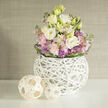 This composition feels like a soft morning in the Chalfonts: layered cream and buttery-yellow lisianthus rise through a halo of blush-pink Stock and petite, spiky purple Allium, all cushioned by leafy green fills. A darker green leaf band encircles the stems at the base, providing a neat, almost corseted finish that draws the eye into the bouquet's centre. The flowers are housed in a remarkable spherical vase-white, sculptural, and constructed from an interlaced web of slender branches that creates a lattice of light and shadow; the vessel is deliberately open so the stems are visible, giving the piece an honest, natural integrity. To the left, two woven wooden orbs-one larger, one smaller-sit on the same white surface, their pale rattan-like strips repeating the vase's woven motif and adding an artisanal rhythm to the tableau. Behind, a muted beige greige wall with a linen or grasscloth texture forms a calm backdrop, letting the pastel tones breathe and the composition feel quietly elegant. I picture placing this arrangement for a home on Gerrards Cross High Street or delivering it to a thoughtful friend near the station; Flower Delivery Gerrards Cross often recommends this palette for sympathy, new homes, or gentle celebrations because its soft hues and tactile materials read as both contemporary and consoling. The scent suggestion is subtle-clean floral musk with a green lift-while the textures invite touch: silky petals, crisp foliage, and the reassuring grain of woven wood. It's a design that brings natural artistry into everyday local interiors without fuss.