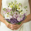 This photograph shows a refined, hand-tied bouquet that seems to glow softly, as if caught in the gentle daylight drifting through a window above Gerrards Cross high street. At first glance, you see a rounded cloud of white lisianthus, their petals layered like fine satin and unfolding in loose spirals that bring an immediate sense of purity and calm. Nestled among them are slender stems of stock in muted lilac and faint blush-pink, each tiny floret adding a subtle wash of colour and a feathery texture that contrasts with the more defined lisianthus blooms. Near the base, a halo of lavender allium forms a playful ring, their tiny star-shaped flowers clustered tightly on rounded heads, adding both structure and quiet drama to the design. Encircling the whole bouquet, ribbons of deep green bear grass have been looped and woven with careful precision, creating delicate arcs and spirals that frame the softer blooms, a touch of modern design that hints at the florist's practiced hand. The palette remains understated and romantic: whites that lean towards cream, lilacs that feel almost silvery, and varying shades of green that recall the tree-lined paths of Bulstrode Park. The flowers appear fresh and cool, as though they have just been lifted from water, droplets still clinging to the stems. You can almost sense the gentle, sweet scent of stock wrapped in the greener, slightly peppery note of allium far beneath. The bouquet's shape is rounded yet not rigid, with an open, breathing top and a more composed, textural base-a design perfect for a thoughtful bride or for someone in Gerrards Cross wanting to send an elegant, hope-filled gesture through Flower Delivery Gerrards Cross.