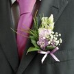 A close-up photograph focuses on the upper body of a man dressed for a formal occasion in Gerrards Cross, his charcoal grey suit jacket filling most of the frame. The fabric of the jacket has a soft, finely textured weave, catching the gentle daylight in a way that highlights the clean line of the lapel and sleeve. Beneath the jacket, a pale dress shirt with a subtle check pattern offers a crisp contrast, while a bold magenta silk tie sits at the centre, its smooth, lustrous surface reflecting a delicate sheen. Pinned neatly to the left lapel is a meticulously crafted buttonhole, its colours and shapes working in quiet harmony. Slender, unopened buds of creamy white flowers rise at the top, reminiscent of delicate snapdragons poised to open. Below them, small blossoms in soft pink and lavender hues cluster together, like sweet peas or miniature stock, their petals gently ruffled and romantic. Tiny white clouds of gypsophila are scattered through the design, adding a light, airy quality that softens the deeper purple florets tucked within. At the base, glossy dark green leaves cradle the blooms, while feathery, brighter green foliage stretches outward with a sense of natural movement, echoing the greenery you might see on a morning walk through West Common. A satin ribbon in mellow lavender is tied into a precise bow around the stems, its sheen echoing the tie's silkiness and giving a refined finishing touch. The overall atmosphere is one of poised celebration, the kind of buttonhole a groom might wear for a church wedding in central Gerrards Cross, radiating joyful anticipation and understated elegance.