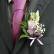 Pinned close to the heart on a dark, finely tailored suit, this boutonnière feels like a quiet promise captured in flowers, the kind often seen at intimate weddings and celebrations across Gerrards Cross. The eye is first drawn to the pale lavender blossoms, their petals soft and velvety, cupping one another in gentle layers that suggest a flower freshly opened in early morning light. Tucked around them are tiny white filler flowers-airy clusters of gypsophila that resemble a light dusting of snowflakes-creating a frothy halo that softens the edges and adds romantic depth. A low collar of glossy green leaves supports the blooms from beneath, each leaf slightly curved, with a well-defined central vein that glints subtly against the dark fabric of the lapel. Finer sprigs of greenery lift upward like playful brushstrokes, giving movement and a touch of wildness, as though the boutonnière has been gathered straight from a spring border in a garden not far from West Common. Around the stems, a sheer lavender ribbon is wrapped and tied with quiet precision; its translucent sheen echoes the colour of the petals and gently mirrors a coordinating tie beneath, suggesting a thoughtfully planned ensemble. The flowers appear fresh and cool, as if they have just been conditioned in clean water at a local studio such as Flower Delivery Gerrards Cross, then arranged with careful hands that understand how to work on this miniature scale. The result is an emblem of grace and romance-perfect for a groom, best man, or special guest-conveying not only style but the tenderness of a day filled with family, friends, and heartfelt vows.