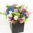 A lush, vibrant bouquet emerges from a modern matte black square box, presenting a vivid display of floristry in the heart of Gerrards Cross. Abundant hydrangeas in periwinkle and indigo provide a plush, textured canvas, intertwined with soft lavender and gentle blush roses, their petals delicately unfurling to reveal layers of colour and emotion. Creamy white lisianthus, punctuated with subtle buttery tones, offer a classic elegance; scattered amidst these are sprigs of whimsical pink astilbe, whose feathery presence lifts the mood with playful grace. Lime-green filler flowers and assorted glossy foliage, reminiscent of the fresh greenery in Gerrards Cross parks, add lively highlights and rich depth throughout. The overall arrangement feels densely abundant yet expertly balanced, perfect for being admired from all sides beneath crisp, natural light. The contrasting black base anchors the composition, bringing focus to the arrangement's radiance and inspiring thoughts of celebration, gratitude, or dignified remembrance. Meticulously crafted for same-day delivery and sophisticated gifting across local Gerrards Cross homes and businesses, this bouquet embodies both contemporary style and the timeless joy of fresh-cut flowers.