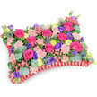 A lovingly crafted rectangular pillow-shaped floral tribute shown at a gentle three-quarter, slightly elevated angle on a clean white background. The surface reads like a plush tapestry: hot pink roses-some tightly furled, others softly unfurling-form the dominant, velvety fields while pale blush-pink carnations weave delicate, ruffled texture between them. Bright lemon-yellow ranunculus or buttercup-like blooms pick out joyful highlights, and deep purple lisianthus punctuate the composition with bell-shaped contrasts that draw the eye inward. Cool, silver-blue eryngium thistles add a hint of architectural spikiness and a wild, maritime coolness against the warmer palette, while rounded silvery-green eucalyptus leaves and deeper green foliage sit underneath like a soft, natural mattress, giving weight and depth. The whole arrangement rests within a richly pleated deep-red satin ribbon that frames the cushion with an elegant, ruffled border and defines the classic pillow silhouette. Soft, diffuse light sculpts each petal and leaf, making the pinks glow and the thistles pick up a subtle steel-blue sheen. The mix of colors-fuchsia, violet, lemon and blush-feels celebratory rather than somber, offering warmth, comfort and a sense of remembered laughter; it is the kind of sympathy pillow designed to honour a life with colour and tenderness. A florist's hand is visible in the tight packing and balanced rhythms of bloom and foliage, a gesture meant to provide solace at service or graveside in Gerrards Cross and neighbouring Chalfont communities.