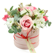 A romantic hatbox bouquet sits perfectly centred against a clear white background, the kind of arrangement you might see prepared early in the morning by a Gerrards Cross florist before deliveries go out across the village. The flowers are gathered into a cylindrical hatbox in the softest pastel pink, its smooth, matte surface lending a contemporary yet graceful feel. A slim coral-red satin ribbon wraps around the box and is tied into a small, precise bow at the front, where a cream tag with flowing green, handwritten-style text hangs lightly, suggesting a thoughtful note or the name of the sender. From the top of the box, a lush dome of blooms rises and spills outward, full but not overcrowded. The eye is drawn first to a large, bi-colour rose in the centre, its pale outer petals gradually deepening to a richer pink swirl in the heart, evoking warmth and affection. Encircling it are white daisy-like chrysanthemums, their crisp petals radiating around bright green centres that bring a lively brightness to the design. Among them, white alstroemeria unfurl with their slender, slightly speckled petals, adding a refined, almost airy detail. Deeper pink and fuchsia-toned flowers, likely spray roses or lisianthus, thread through the softer hues, adding pockets of intense colour that keep the arrangement dynamic. A blend of greenery, including rounded silver-green eucalyptus leaves, deeper green filler foliage, and delicate stems tipped with tiny buds, creates depth and texture, softening the outline of the bouquet. The composition feels fresh, joyful, and considered, perfectly suited to anniversaries, birthdays, Mother's Day, or a romantic surprise delivered to a doorstep near West Common, capturing the gentle, elegant spirit of Gerrards Cross in floral form.