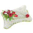 A solemn, carefully crafted rectangular sympathy pillow sits portrayed against a soft, neutral background, its entire surface densely carpeted with hundreds of small, pristine white chrysanthemum blooms that read like a cushion of fresh snow. The chrysanthemums form a velvety, cloud-like texture that suggests quiet rest and reverence, while a slim border of pale green satin ribbon or foliage peeks out along the slightly raised edge, giving the piece a gentle, framed finish. On the top left corner an articulate spray of deep red blooms anchors the design: velvety red roses in various stages from bud to open bloom, paired with richly hued red carnations whose ruffled petals add shadow and depth. Interspersed among them are tiny white accent flowers-likely freesias or spray chrysanthemums-that introduce delicate highlights and a whisper of sweetness. Lush green foliage, from feathery fern fronds to broader glossy leaves, provides a verdant backdrop and lifts the reds from the white field. Across the white surface, three slender arcs of bright green bear-grass or similar linear foliage sweep diagonally toward the lower right, drawing the eye and imparting a graceful motion that culminates in a single, perfectly formed red rose placed as a poignant focal point. The composition is softly lit, carrying a faint, clean floral scent, and the overall mood is one of dignified remembrance. As a local florist would note, this traditional cushion is appropriate for funerals, memorial services, or as a comforting tribute delivered to churches and funeral homes around Gerrards Cross and neighbouring villages, offering an understated expression of love, honour, and solace.