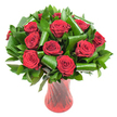 A luxurious cluster of roughly a dozen deep crimson roses fills the frame, each bloom in full, velvety perfection with petals spiralling from a tight centre into plush, layered cups. The colour reads almost wine-dark in the soft, even studio light, which brings out the subtle sheen on each petal and the delicate shadow between the layers. Surrounding and lifting the rose heads is an abundance of rich, dark green foliage: glossy, flat leaves that form a dense verdant backdrop, likely Salal or Italian Ruscus, and larger, brighter emerald leaves-suggestive of Aspidistra-that have been carefully curled and rolled into sculptural loops. Those folded leaves create graceful arcs that cradle the blooms and add a modern, architectural rhythm against the softness of the roses. The bouquet sits in a tall, slender ruby-red glass vase with a faint ribbed twist that catches the light in vertical highlights, its translucent depth echoing the intensity of the roses without overpowering them. Set against a pure white background, the reds and greens pop with clear contrast, while the composition's clean lines and generous volume communicate both celebration and quiet refinement. There's an implied fragrance here-classic, heady rose scent-and a tactile invitation to brush a fingertip along the velvety petals. This arrangement, reminiscent of pieces I prepare for anniversaries or a thoughtful Valentine's delivery around Gerrards Cross High Street and the station area, reads as an elegant, heartfelt statement; it feels engineered to be both a romantic gesture and a polished centrepiece for a dining table or mantle.