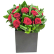 A study in modern romance: a compact cluster of deep crimson roses sits at the heart of a low, square matte-black box, each bloom opening in velvety spirals that catch the light and hold the eye. Around them, broad glossy leaves have been carefully curled and overlapped to form a rhythmic, almost architectural frame; the foliage's vibrant green gives the composition a sculptural depth and a sense of movement, like waves frozen mid-swell. The lighting is crisp and even, lifting highlights from the petals and the sheen of the leaves so the crimson reads as both rich and distinctly velvet-soft, while the greens range from bright emerald to jade where shadows fall. Texturally the contrast is delicious: the silken, yielding petals against the firmer, waxy curves of the greenery, and the matte box base that anchors the design with a contemporary, urban edge. There is a subtle perfume on the breeze - the classic, heady sweetness of rose tempered by the fresh, green tang of newly cut leaves - that suggests a moment of celebration or a quiet declaration of affection. As a local florist speaking from Gerrards Cross, I imagine this arrangement gracing a sophisticated dining table on the High Street or arriving as a thoughtful anniversary gesture for someone returning home from Gerrards Cross station; it's equally at ease in a modern office or a cosy sitting room. Every fold of leaf and placement of bloom feels intentional, designed to make a lasting visual and sensory impression without fuss, a bold yet refined expression of love and admiration.