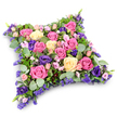 A beautifully detailed cushion-shaped funeral tribute is pictured against a pure white background, the floral design forming a soft, padded rectangle that calls to mind a comforting pillow laid gently beside a casket or memorial in Gerrards Cross. The arrangement is tightly composed so that the entire surface is a continuous tapestry of blooms, with no visible gaps. Rich pink roses in shades from deep fuchsia to gentle blush sit alongside creamy, pale yellow roses, each blossom at a slightly different stage of opening, creating a natural rhythm of tighter buds and fully open, layered rosettes. Nestled among them are elegant lisianthus flowers in deep royal purple and velvety violet, their softly folded petals and rounded shapes introducing both depth and a sense of quiet luxury. Between these focal flowers, clusters of tiny lavender and lilac filler blooms, likely statice or limonium, add a misty, textural quality, like a delicate veil laid across the petals. A generous collar of greenery frames and supports the cushion: rounded eucalyptus leaves in soft, silvery green mingle with darker, finer foliage reminiscent of ferns, together lending structure and a calm, natural backdrop to the brighter colours. The lighting is even and gentle, creating subtle shadows that define the flower heads and accentuate the cushion's gently rounded edges. The mood conveyed is tender, reflective, and deeply respectful, ideal for a final tribute from close family or friends. This floral pillow would feel perfectly at home at a service in one of Gerrards Cross's churches or as a graveside offering at the local cemetery, a quiet yet eloquent way to say goodbye and express enduring love.