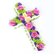 A traditional floral cross is shown resting against a clean white background, positioned diagonally so that the top left appears gently raised, lending the tribute a sense of quiet motion. The entire silhouette of the cross is edged with compact white spray chrysanthemums, their small petals tightly layered to form a smooth, cloud-like frame. Just beneath this soft border, a band of light green ribbon or folded foliage subtly circles the perimeter, adding a polished finish and a fresh hint of colour that cradles the flowers within. The interior of the cross is lavishly filled with an artful mix of blooms: fully opened roses in vivid fuchsia pink and soft, buttery cream draw the eye along the arms and centre, their velvety petals suggesting tenderness and love. Interspersed with these larger roses are bright lime-green button chrysanthemums, their rounded, textured heads giving a lively contrast that keeps the design from feeling heavy. Smaller blossoms in deep purple and violet, likely statice or lisianthus, weave through the gaps, bringing depth, richness, and a sense of quiet reflection. Lit with clear, even light, each colour-from the crisp white edge to the jewel-toned centre-is clearly defined, much as it would appear laid before the altar of a church or chapel in Gerrards Cross. The overall mood is one of respectful remembrance, with the cross shape gently affirming Christian faith while the vibrant palette celebrates the life of the person being honoured. It is an ideal floral tribute for funerals, memorial services, or graveside tributes in and around Gerrards Cross.