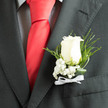 In the soft, directional light of an arrival hall, an ivory rose boutonnière stands in crisp contrast against a smart charcoal jacket, its velvety contours echoing the tailored lines of a checked shirt and the decisive flash of a red silk tie. The rose's petals unfurl with a poised, velveteen texture that suggests steadfastness and tenderness at once; clustered at its base, the baby's breath offers a veil of tiny white blossoms that read as purity and quiet devotion. Slender blades of fern and gently twisted leaves arc around the bloom, their deep green hues and glossy surfaces creating movement and sculptural depth within this otherwise minimalist piece. A fine white ribbon, tied with firm, practiced hands, secures the assembly and adds a ceremonial whisper against the darker fabrics. You can almost sense the cool, fresh touch of the flowers and the faint, classic scent of rose that lifts when the wearer turns-an evocative detail for an early summer wedding or a formal celebration at one of Gerrards Cross's parishes or local halls. As a local florist arranging buttonholes for clients arriving from Chalfont Road or the station, I would balance durability with softness so the boutonnière travels well yet reads as immediate and alive next to a red tie or check-pattern shirt. For those who order through Flower Delivery Gerrards Cross, this is the little emblem we prepare with attention to proportion, ensuring it complements both the outfit and the significance of the day without competing with it.