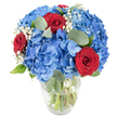 A fresh, celebratory bouquet sits in a simple clear glass vase, lit by soft daylight that brings the saturated colours into bright relief. The composition is built around lush, rounded clusters of sky-blue hydrangeas whose many small petals overlap to form cloud-like domes; their cool, watery blue anchors the arrangement with a serene, summery feeling. Nestled among the hydrangea masses are three deep red roses, their velvety petals still tightly spiralled, providing a passionate counterpoint that draws the eye. Fine spray of white baby's breath threads through the blooms like drifting clouds, its tiny florets adding an airy punctuation and a sense of lightness. Muted green eucalyptus leaves, oval and slightly silvered, weave through the bouquet, offering soft, textured contrast and the faint herbal note that lifts the bouquet's scent. The vase's gently rounded base and tapered neck reveal green stems crisscrossing beneath the water line, a clear sign of freshness and careful arranging. Set against a clean white background, each colour and texture reads clearly-the cool blue, the warm red, the crisp white, the calming green-so the arrangement feels both modern and comforting. As a local florist from the Gerrards Cross area might suggest, picture this on a kitchen table near the station after a market run, bringing a quiet but confident joy to a birthday, anniversary, or relaxed patriotic gathering. The bouquet's balanced contrast of hues and textures makes it equally suited to brighten a living room or to be given as a heartfelt, elegant gift.