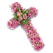 A deeply expressive floral cross stands alone against a bright, uncluttered white background, its entire structure transformed into a tapestry of pink and mauve blossoms. The shape of the cross is clear yet softened by the abundance of flowers, giving the impression of a cushioned, petal-covered form that feels both gentle and substantial. At the centre point, where the vertical and horizontal arms meet, three fully open roses in a muted dusty rose and pale mauve hue form a calm, central focus; their softly spiralling petals seem to cradle the emotions of farewell and gratitude. Around them, slender fern fronds and sprigs of fresh green foliage weave between petals, adding natural contrast and a touch of garden-like freshness to the design. The rest of the cross is richly filled with smaller lisianthus blooms in varying shades of soft pink, brighter rose, and delicate lavender, many with characteristic darker centres that pull the gaze inward. Intermingled clusters of tiny pink and purple filler flowers, along with specks of pale green buds and petite white blossoms, create a finely textured surface that feels intricate yet harmonious. Along the very edge of the cross's outline, a narrow hint of darker purple ribbon or fabric base peeks through, offering a discreet frame that enhances the floral shape without overpowering it. The overall mood is tranquil and reverent, a traditional Christian symbol rendered in soft, comforting tones. It evokes the kind of floral tribute chosen in Gerrards Cross for meaningful funeral services and memorial gatherings, conveying respect, enduring love, and quiet spiritual hope for the person being remembered.