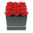 A meticulously arranged display of preserved red roses is shown in a close, eye-level view, the style of design often requested for romantic gestures across Gerrards Cross. Twelve roses, each one fully open and nearly identical in diameter, are nestled tightly together in three neat rows of four, covering every inch of the box's top. Their petals are lush and layered, forming deep, velvety spirals where a rich ruby red shade softens into darker crimson tones in the inner curls. The blooms rise slightly above the rim of a square, matte black box, giving the impression that the flowers are gently overflowing without losing their precise structure. The box's sides are smooth and untextured, with sharp, architecturally clean corners that lend a contemporary, almost sculptural quality to the arrangement. Around the lower section runs a slim white stripe, a crisp horizontal detail that adds refinement without distracting from the roses themselves. Against a pure white background, the contrast of black, red, and white feels dramatic yet balanced, reminiscent of the polished style favoured in modern homes near West Common and the leafy streets leading out of Gerrards Cross. Soft, even lighting picks out the curves of each petal, creating subtle shadows that enhance the sense of depth without any harsh edges. There are no visible stems, leaves, or decorative extras, just the pure, distilled idea of romantic luxury in floral form. The image evokes anniversaries, Valentine's surprises, and intimate celebrations at home, suggesting a premium rose box delivered to mark a moment of sincere affection in Gerrards Cross.