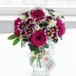 A vibrant mixed bouquet of fresh flowers stands proudly in a clear, textured glass vase, its rounded shape cradling a joyful explosion of pinks, purples, whites, and deep greens. At the heart of the arrangement, large gerbera daisies in rich fuchsia and magenta draw the eye, their velvety petals radiating from dark central disks that add depth and drama. Around them cluster bicolour chrysanthemums, their many fine petals tipped in deep purple and softening to white as they reach the centre, where bright yellow eyes seem to sparkle in the light. Creamy white roses, some fully open and others still in tight, hopeful buds, thread through the design, lending an elegant, timeless note that softens the bold colour palette. Delicate sprays of white gypsophila float between the larger blooms like tiny clouds, giving the bouquet a light, airy texture that contrasts beautifully with the plush petals and glossy foliage. Deep green leaves frame the flowers and gather at the base, where their stems are visible through the water-filled vase. A simple band of rustic twine is tied neatly around the neck of the vase, finished with a bow from which a small white gift tag hangs, decorated with a subtle pink floral emblem and soft lettering, suggestive of a personal message for a birthday, anniversary, or thank you. The background is a softly diffused, bright white, reminiscent of daylight streaming through a Gerrards Cross window, allowing the colours to glow and making the arrangement feel freshly delivered, as if it has just arrived on a hall table off Packhorse Road or a sunny kitchen counter near East Common. The overall impression is of joy, care, and celebration, captured in flowers that seem ready to brighten any home in Gerrards Cross.
