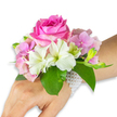 A delicate wrist corsage captured against a clean white backdrop, shown on a left hand and forearm with a light-to-medium skin tone, as if ready to be worn for a first dance or wedding vow. The focal bloom is a large, fully opened deep fuchsia-pink rose whose tightly layered petals form a plush spiral; its velvety surface catches a soft, natural light that suggests the faintest perfume of rose. Around it, clusters of blush-pink hydrangea florets create a cloud-like volume, each tiny petal slightly translucent and providing a gentle cushion for the composition. Pristine white alstroemeria (Peruvian lilies) sit at intervals, their four-to-six petals edged with a whisper of pale green streaking toward the throat, lending crisp contrast and a hint of botanical geometry. On the right side of the corsage a small spray of light green alstroemeria buds and a few barely open pink buds promise further bloom, their taut forms adding movement and anticipation. Broad, glossy emerald-green leaves cradle the flowers, their rich tone balancing the pastels and intensifying the overall colour. The arrangement is secured to the wrist by a slender, multi-strand bracelet of small lustrous white pearls that loop around the wrist and trail slightly down the back of the hand, catching light with a soft sheen. The composition reads as a wearable bouquet-refined, fragrant, and youthful-perfect for prom, bridesmaids, or a celebratory gathering in Gerrards Cross, where shoppers on the High Street often choose this classic, hand-crafted style for special occasions.