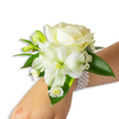 A delicate wrist corsage captured against a soft white backdrop, composed with the quiet grace of a seasoned florist's hand. At its center sits a creamy ivory rose, its velvety petals unfurling in layered curls that catch the light with a warm, soft sheen. Nestled just below and slightly left is a larger, trumpet-shaped white bloom-an alstroemeria-like flower with a subtle lime-green throat that adds a luminous depth and a gentle contrast to the rose's cream. Around these anchors, tiny cloud-like sprigs of Gypsophila breathe airiness into the design while small, bright white daisy-style florets with sunny yellow centres lend a playful, tender detail near the base. Several fresh green buds, still tightly closed, promise movement and life as if the corsage will open a little more through the evening. Broad, glossy deep-green leaves frame and cradle the arrangement, their smooth waxy texture providing a lush backdrop that amplifies the purity of the whites. The whole piece is secured to the wrist with a multi-strand pearl bracelet: lustrous, round faux pearls sit cool and smooth against fair skin, the strands looping with gentle elegance and lending the piece a classic, tactile luxury. In soft morning or gallery light this corsage conjures notes of powdery freshness and gentle perfume-a scent memory of spring gardens. Designed with ceremony in mind, it evokes weddings, proms, and milestone celebrations in Gerrards Cross, where local brides and families often seek that quiet, polished charm for their most cherished moments.