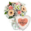 Photographed against a clean white background, this Mother's Day arrangement looks like it has just been set down on a hall table, ready to greet a mum as she walks through the door in Gerrards Cross. A clear, subtly shaped glass vase holds a full, rounded bouquet of mixed flowers, the glass tied at the neck with a simple loop of light brown twine that adds a homely, handcrafted touch. The bouquet is a gentle blend of soft spring colours: several peach-pink roses are in full bloom, their velvety petals unfurling in classic spirals that feel luxurious yet comforting. Around them, peach gerbera daisies with deep, dark centres bring a modern, graphic note, their wide, even petals radiating out like spokes on a wheel. Sprays of delicate white baby's breath thread through the bouquet, their tiny blossoms forming a lacy veil that lightens the whole design, while small white and pale green chrysanthemums tuck into the spaces, giving extra volume and a crisp, fresh energy. Glossy green foliage frames the flowers and spills slightly over the rim of the vase, suggesting abundance without feeling untidy. To the right of the bouquet, attached but slightly in front, floats a heart-shaped Mylar balloon. Its surface is a bright white or soft cream, scattered with playful golden-peach and metallic dots that catch the light like confetti, and at its centre a peach heart encloses the words Happy Mother's Day in flowing, coral-peach script. Just visible between the vase and the balloon is the edge of a small white greeting card, hinting at a handwritten message inside. The whole scene conveys warmth, gratitude, and gentle celebration, the kind of carefully chosen gift local customers might order for same-day delivery in and around Gerrards Cross.