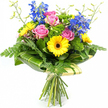 Captured against a bright white background, this colourful hand-tied bouquet appears as if it has just been lifted from a florist's workbench in Gerrards Cross, dewy and brimming with life. At the centre, soft pink roses open in generous spirals, each petal gently curling back to reveal the heart of the bloom, conveying warmth and affection. Around them, bright yellow gerbera daisies with striking dark centres add an unmistakable burst of cheer, their flat, open faces giving the arrangement a friendly, upbeat character that would brighten any living room near East Common or a desk in a local Gerrards Cross office. Towering above the main cluster, deep blue delphinium stems introduce graceful height and a serene, cool contrast, the individual florets layered along each spike like tiny bells. The bouquet is wrapped in an abundance of greenery, from fine, feathery fern fronds softening the outer edges to broad, glossy green leaves shaped into an elegant collar around the lower sections of the blooms. Some of these larger leaves show subtle lighter green stripes, adding quiet visual interest and a hint of tropical flair. Between the feature flowers, bright green textured filler blooms and foliage create a lush, almost moss-like underlayer that enhances the sense of depth and freshness. Slender, light green stems with small buds stretch gently above the main flowers, giving an impression of movement and new life. At the base, the natural stems are gathered tightly and tied with a simple bow of ribbon or twine, emphasising the handcrafted nature of the design. The whole bouquet feels joyful and uplifting, ideal for same-day delivery around Gerrards Cross to celebrate birthdays, anniversaries, or spontaneous moments of appreciation.